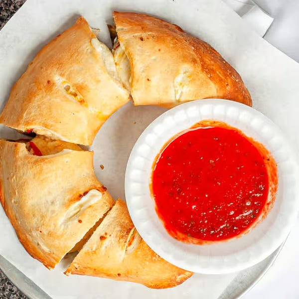 A golden-brown calzone, sliced into pieces, with a side of red marinara sauce in a white bowl.