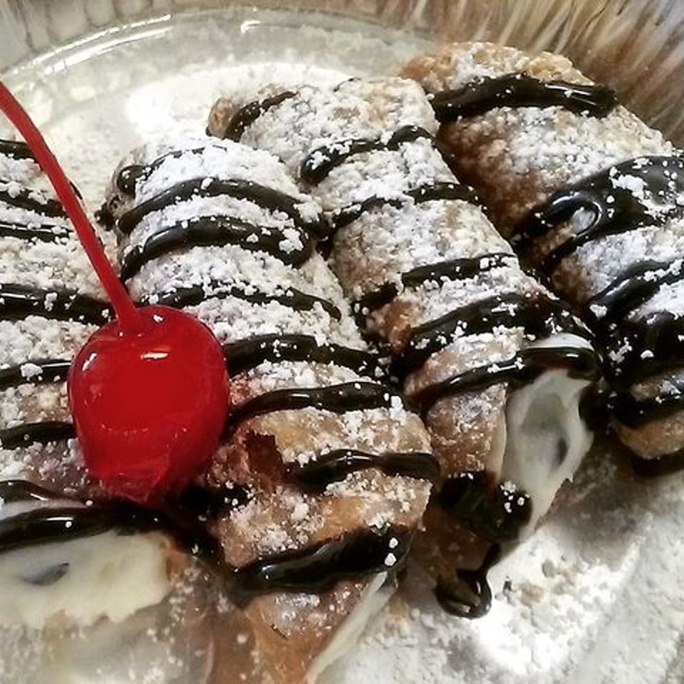 Four cannolis drizzled with chocolate and powdered sugar, garnished with a red cherry.