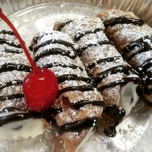 Four cannolis, dusted with powdered sugar and drizzled with chocolate, garnished with a bright red cherry.